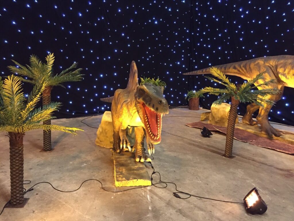 a dinosaur model in a room with palm trees and lights