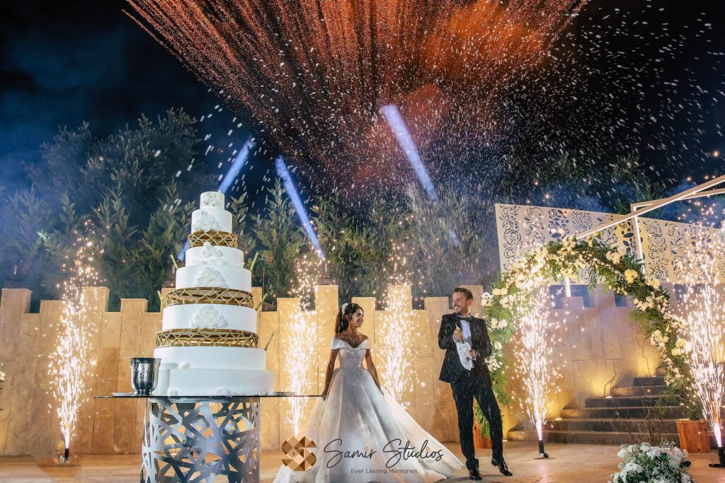 a couple in wedding attire standing next to a large cake with fireworks