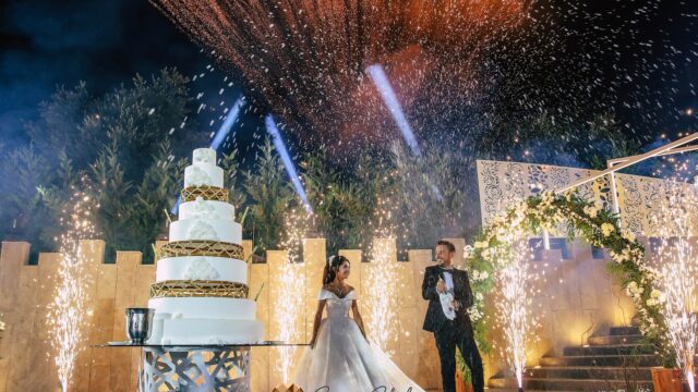 a couple in wedding attire standing next to a large cake with fireworks