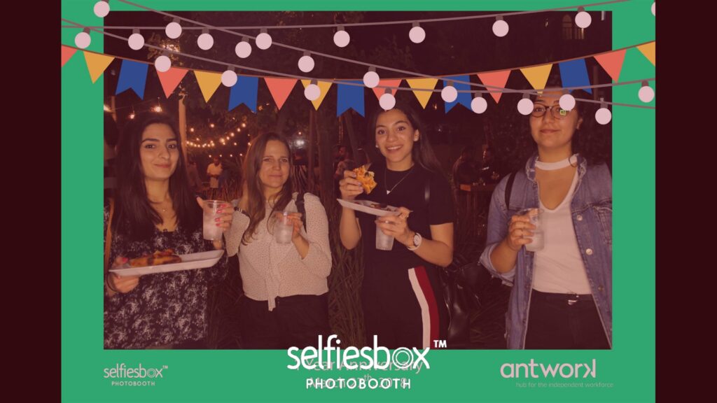 A group of women holding drinks and standing under string lights at the "Selfiesbox Photobooth."