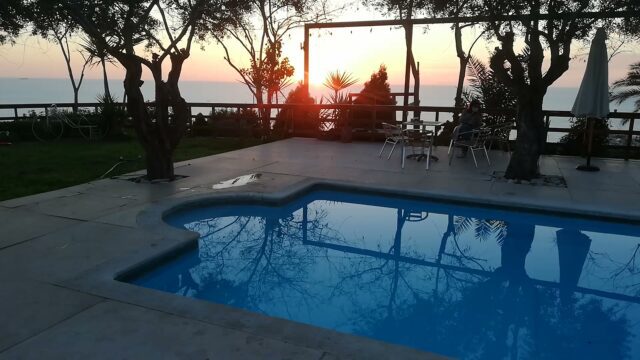 a pool with a table and chairs by a deck with trees and a sunset