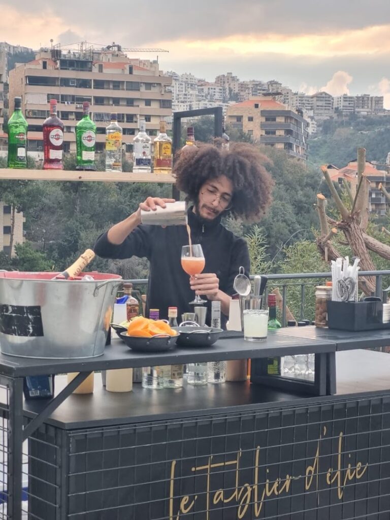 A man, wearing a "Le Tablier D'elie," is pouring a drink into a glass.